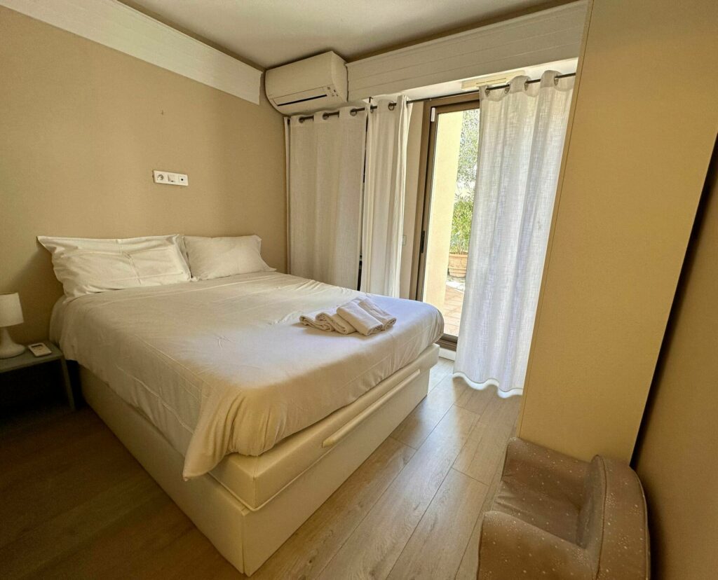 Chambre double avec lit blanc, baie vitrée, fauteuil en cuir, tons beiges et dorés – appartement Cap d'Ail